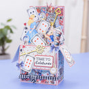 A decorative 3D pop-up card box themed like the Alice in Wonderland 'Time to Celebrate' scene, featuring playing cards, a golden key, a pocket watch, and a checkered ribbon on a light blue surface.