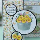 A handmade greeting card with a floral patterned background, featuring layered circular cutouts with yellow sunflowers, pink blossoms, butterflies, and a galvanized bucket. One circular label reads 'Have a beautiful BIRTHDAY'.