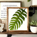 Framed green leaf print on a shelf with a book and plant in the background