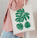 A person wearing a pink corduroy jacket and light blue jeans carries a white canvas tote bag with green tropical monstera leaf prints.