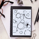 A spiral-bound notebook with a black cover and a white page featuring black ink-style circle and splatter drawings, photographed on a light textured surface with a few office props around it.