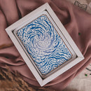 A square white-framed art print featuring a blue and white swirling pattern, styled on draped dusty pink fabric with dried grass accents.