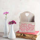 A rustic wooden magazine holder with a heart-shaped cutout at the top and a pink floral stencil design on the front, styled on top of two books next to two white vases with pink flowers against a textured light gray wall.