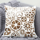 A square decorative throw pillow with a white background and laser-cut brown gear and cog pattern, resting on a gray knit blanket.