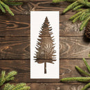A rectangular white stencil or template with a laser-cut pine tree design, displayed flat on a dark rustic wooden background with green pine branches and a pinecone.