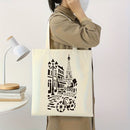 A person wearing a beige shirt and white face mask carries a cream canvas tote bag with a black Paris-themed graphic print featuring the Eiffel Tower, street lamps, and a bicycle. The background is a clean white wall with a white desk and lamp on the left.