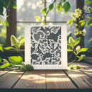 A square white art print or card featuring an intricate black lace-like floral and scroll pattern, displayed on a wooden surface surrounded by green leaves and small white blossoms in a sunlit outdoor setting.