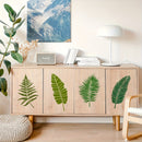 A light wood sideboard cabinet with four large doors decorated with green tropical leaf decals, shown in a styled room setting with a lamp, books, a speaker, and a plant.