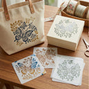 A canvas tote bag and matching gift box with butterfly and floral stencils, displayed on a wooden table alongside stencil sheets, ribbon spools, and scissors.