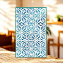 A rectangular decorative panel with a repeating blue leaf or snowflake-like geometric pattern on a white background, standing upright on a wooden table in a softly blurred indoor setting.