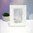 A white square art print or card featuring a purple abstract design of vertical streaks and small star-like marks, displayed upright on a white surface with a small potted succulent and a soft purple bokeh background.