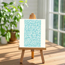 A small wooden easel stands on a sunlit wooden table, holding a white art print with a light blue abstract pattern. The background shows a softly blurred indoor setting with a window and green potted plants.