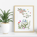 A framed botanical art print featuring colorful flowers, green leaves, and small birds, styled on a white shelf next to a potted green plant in a white pot with gold trim.