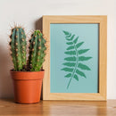 A framed botanical art print featuring a green fern leaf on a light blue background, styled next to a potted cactus on a wooden surface against a neutral wall.