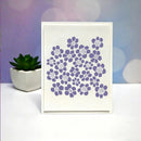 A square white card featuring a cluster of purple daisy-like floral cutouts, displayed on a white surface with a small potted succulent and a soft purple-blue bokeh background.