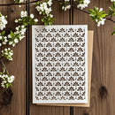 A square white card with an all-over repeating leaf cutout pattern, placed on top of a kraft paper envelope on a rustic wooden surface with white blossoms around it.