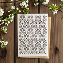 A white rectangular stencil sheet with an ornate repeating floral cutout pattern, placed on top of a tan envelope on a rustic wooden surface with white blossoms around it.