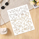 A white rectangular card or stencil with an intricate floral and vine cutout pattern, laid flat on a light wood surface with a beige woven textile, sunglasses, a small cream purse, and pale flowers nearby.