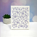 A square white card with a delicate purple floral vine pattern, standing upright on a white surface with a small potted succulent and a soft purple bokeh background.
