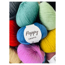 A close-up arrangement of colorful yarn skeins with a white paper label reading 'Poppy CRAFTS' in the center. The visible yarn colors include teal, mint green, blue, red, yellow, pink, beige, and dark gray.