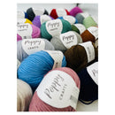A close-up arrangement of multiple yarn skeins in assorted colors, each with a white paper label reading Poppy Crafts. The visible colors include pink, blue, teal, gray, brown, navy, purple, green, and cream, photographed on a light neutral background.