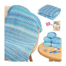 A crocheted blanket in variegated light blue, aqua, and white yarn draped over a chair, with folded matching yarn shown at top right and a round wooden table at bottom right holding several blue yarn balls, crochet hooks, needles, and tools. A small video icon and booklet are also visible.