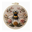 A circular embroidery hoop containing a detailed floral and insect textile design, featuring a bumblebee at the center surrounded by pink flowers, green leaves, and gold-toned botanical elements on a light beige fabric background.