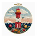 A circular embroidery hoop containing a finished embroidered scene of a red and white lighthouse on a rocky shore, with blue sky, clouds, flowers, and dark blue hills. The hoop is centered on a plain white background.