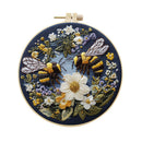 A circular embroidered needlework piece in a wooden hoop, featuring two bumblebees and white, yellow, and purple wildflowers on a dark navy blue background.