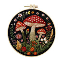 A circular embroidery hoop containing a detailed floral and mushroom embroidery design on a black fabric background. The design features three large red-capped mushrooms with white spots, surrounded by green leaves, small flowers, and wild grasses in red, orange, pink, yellow, and white.