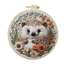 A circular embroidery hoop holding a detailed stitched hedgehog surrounded by small flowers and leaves on a white background.