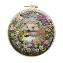 A circular embroidery hoop displaying a detailed stitched artwork of a brown and cream hedgehog surrounded by colorful wildflowers on a pale blue-green background, shown against a plain white backdrop.