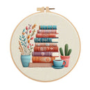 A circular embroidery hoop containing a stitched illustration of stacked vintage books, a potted plant, a cactus, and a blue cup on a light fabric background.
