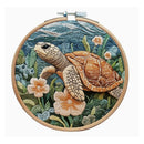 A circular wooden embroidery hoop holding a detailed needlepoint or embroidered sea turtle scene with blue-green water, green plants, and peach flowers, shown against a plain white background.