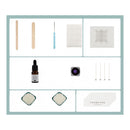 A flat-lay collage of craft or DIY kit components on a white background with a light blue border. Items include wooden sticks, a blue-handled tool, a square white sheet, a translucent square piece, a small amber dropper bottle, a round purple-tinted square, four thin metal rods, two powder samples, and a card reading THANKYOU.