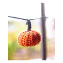 A small orange crocheted pumpkin ornament hanging from a black string against a softly blurred window background.