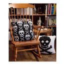 Two crocheted throw pillows with black and white skull patterns are displayed in a cozy, rustic room setting. One pillow is propped up on a wooden rocking chair, while the other lies on the floor next to it.