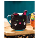 A black crocheted cat-shaped mug cozy with pink ears, green eyes, and white whiskers, shown wrapped around a blue mug on a wooden tray.