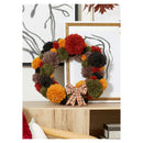 An autumn-themed pom-pom wreath with orange, red, green, brown, and black pompoms, decorated with a plaid bow, displayed on a wooden shelf in front of framed wall art.