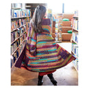 A woman viewed from behind wearing a large, hand-crocheted striped blanket or shawl in a library or bookstore aisle, surrounded by bookshelves.