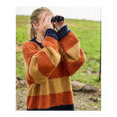 A woman outdoors wearing a chunky knit sweater with wide horizontal stripes in rust orange, tan, and navy blue, holding black binoculars to her eyes. The background shows a soft-focus grassy field.