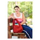 A smiling woman sits on a wooden bench outdoors, holding a bright red chunky-knit shoulder bag. She is wearing a white tank top with pastel horizontal stripes and navy blue track pants with a white side stripe.