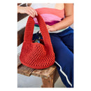 A person sitting on a wooden bench holds a crocheted shoulder bag in a warm rust-red color. The person is wearing a pink and purple striped tank top and blue sweatpants with a white stripe.