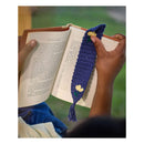 A navy blue crocheted book bookmark with yellow and green flower details is held over an open book by two hands, with a blurred outdoor background.
