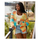 A woman wearing a colorful, open-front crochet cardigan with stripes of yellow, orange, pink, and blue, paired with a white tank top and denim shorts, standing outdoors near a railing.