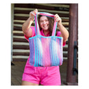 A smiling woman holds up a large woven tote bag with vertical pink, blue, and purple stripes. She is wearing a pink shirt and shorts, standing in front of a rustic wooden log building.