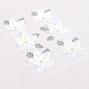 White paper flower stickers with gold leaf vein details and small green leafy sprigs, arranged on a clear sheet over a light beige background.