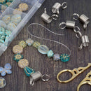 Jewelry making supplies arranged on a dark wood surface, including silver coil end pieces, clear wire, faceted beads in turquoise, cream, and gold, round embellishments, a clear compartment box of beads, and gold-toned scissors.