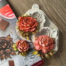 A white silicone mold tray with floral cavities, shown alongside three 3D-printed peony flower models in coral, pink, and peach tones with green and yellow leaves. The scene is styled on a wood-grain tabletop with craft-related props, including a red palette and wooden blocks.