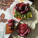 Three carved rose sculptures in shades of deep burgundy and purple, displayed alongside a white floral mold and a wooden carving block on a split white and gray wooden background.
